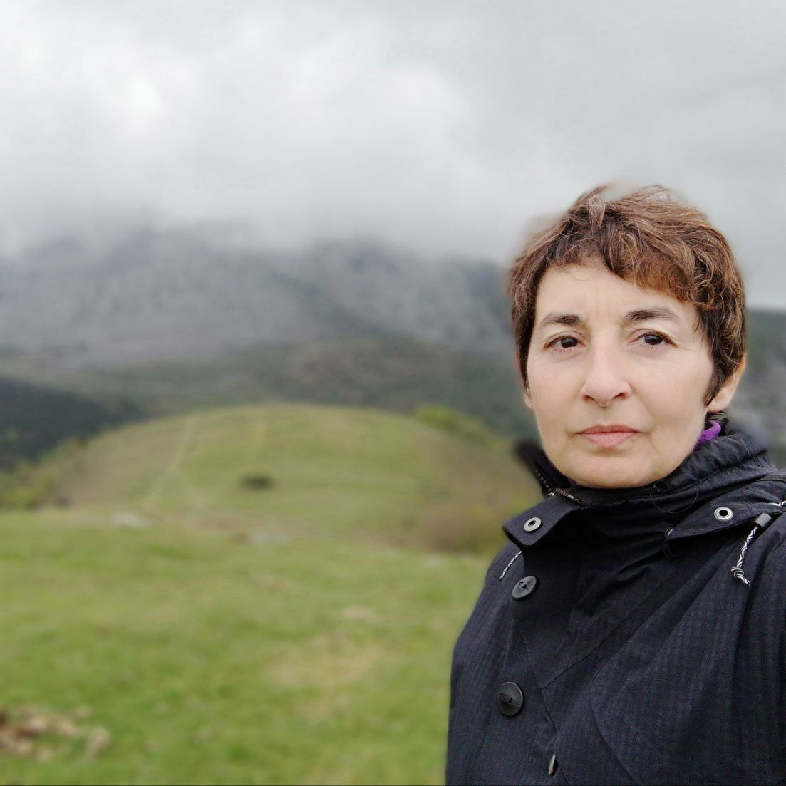 Se ve a Ana mirando a cámara, y se ve que está en el campo con montañas altas, y nubes. Está abrigada